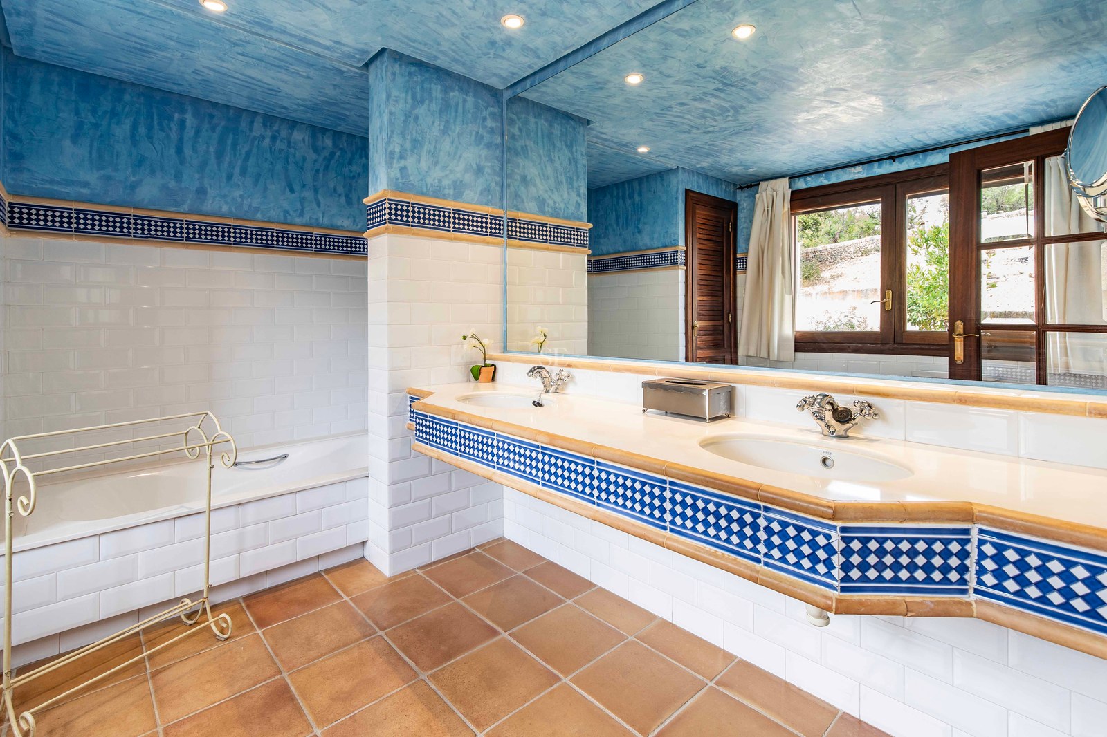 Salle de bain avec plafond bleu texturé, sol en terre cuite, double vasque et baignoire encastrée.