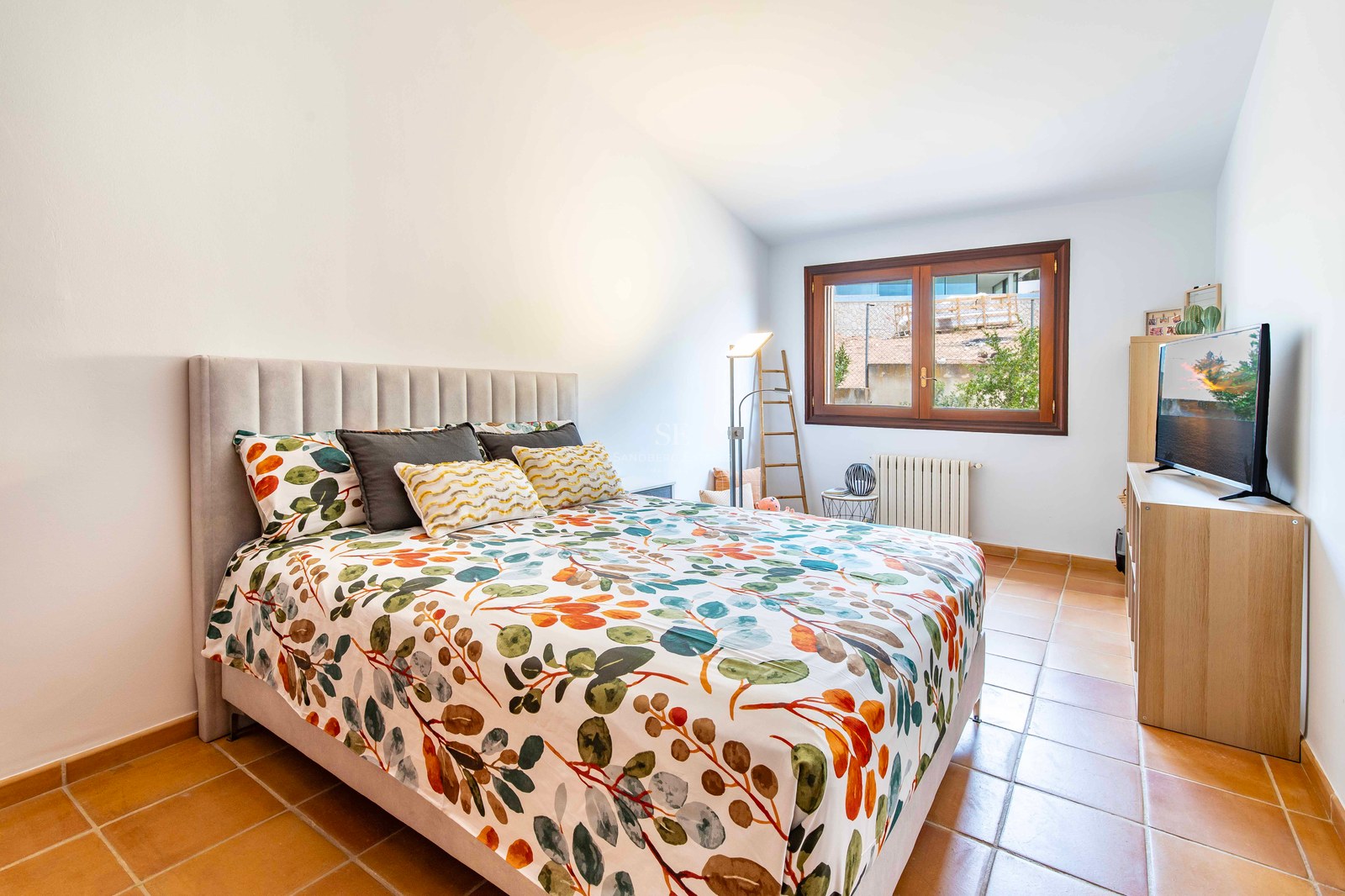 Chambre avec lit à motifs floraux, tête de lit grise, sol en terre cuite et fenêtre en bois.