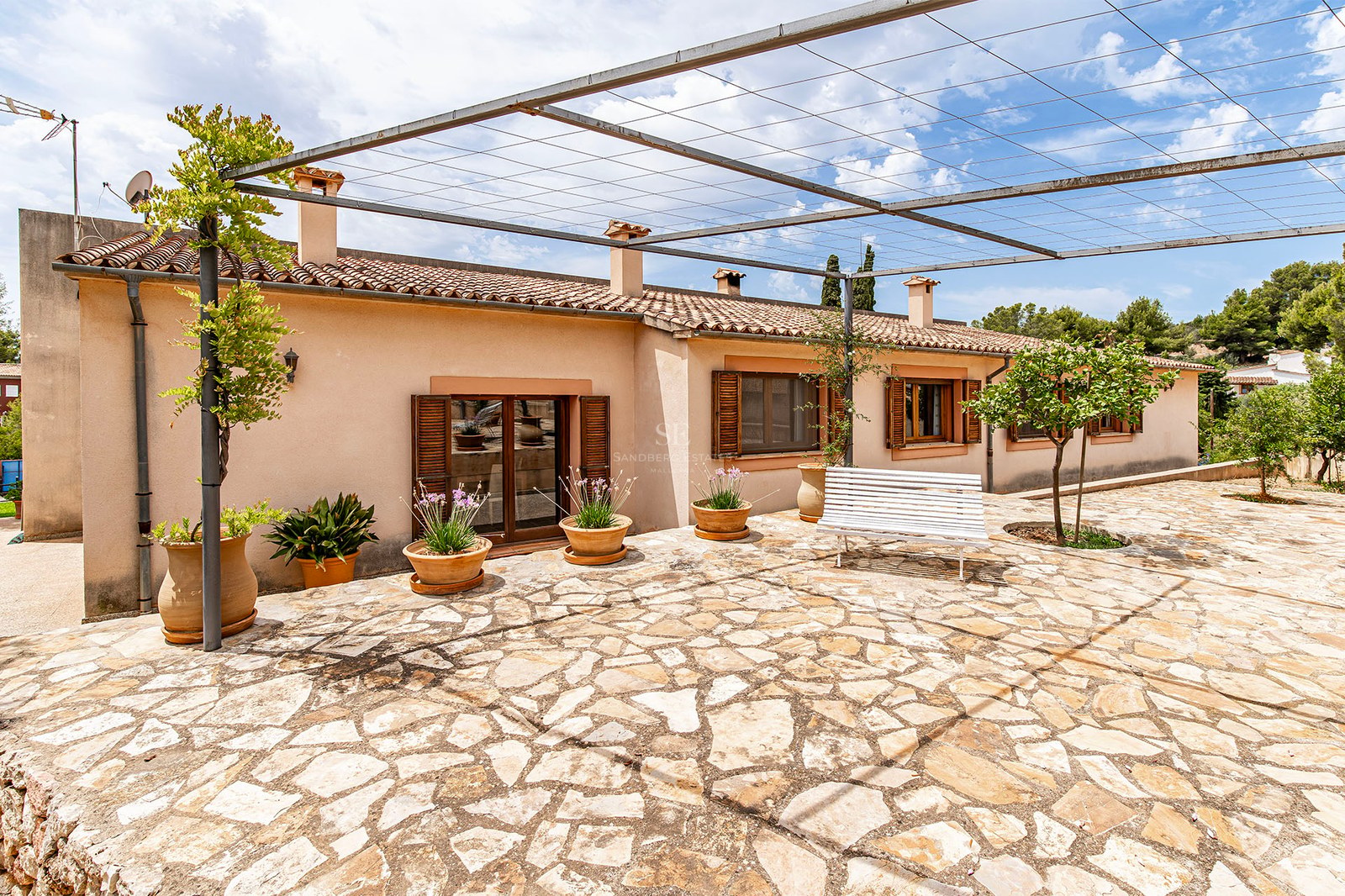 Terrasse pavée de pierre avec pots en terre cuite, pergola métallique et banc blanc devant une maison rustique.