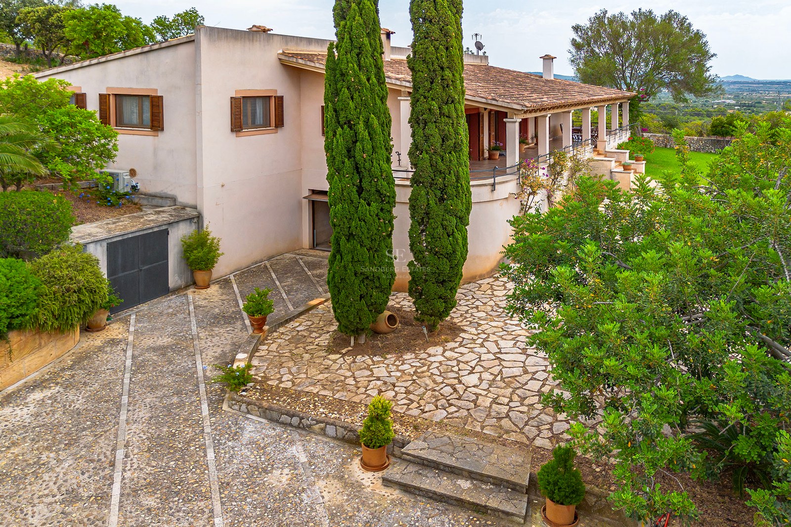 Vue d'une villa méditerranéenne aux murs roses avec une cour en pierre naturelle et de grands cyprès.