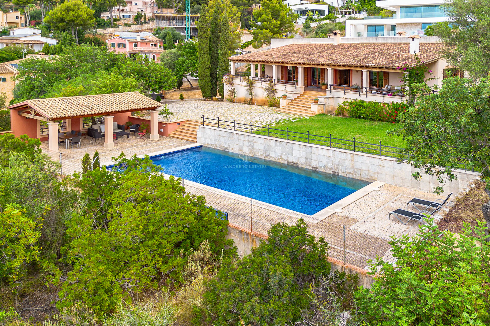 Vue aérienne d'une grande piscine rectangulaire bleue entourée de dalles en pierre, un pool house et une villa méditerranéenne.