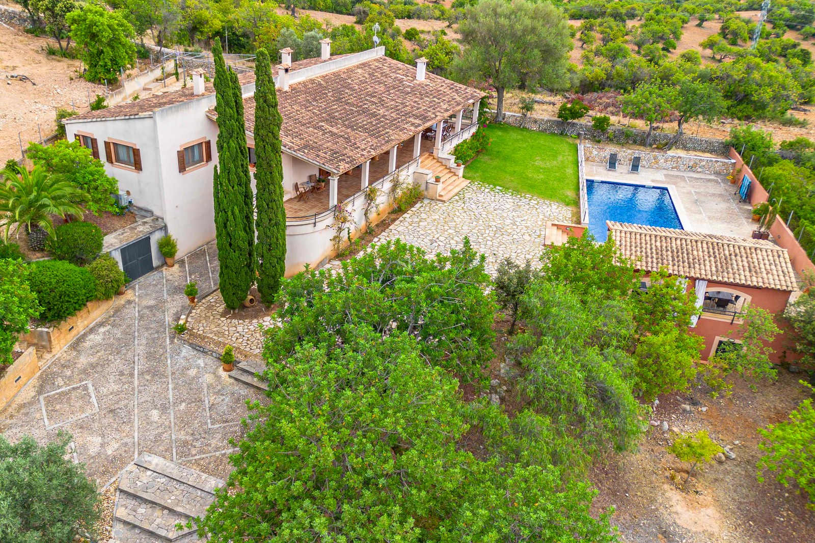 Vue en plongée d'une villa avec toit en terre cuite, cour en pierre, piscine bleue et aménagement paysager verdoyant.