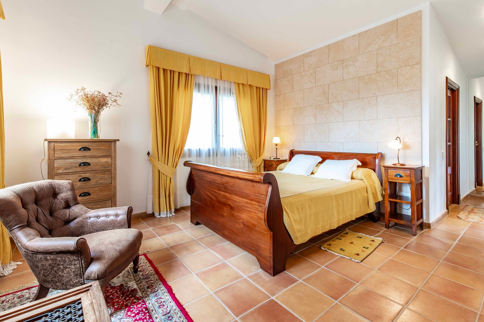 Une chambre rustique avec un lit en bois, des tomettes en terre cuite, un fauteuil en cuir et des rideaux jaunes.