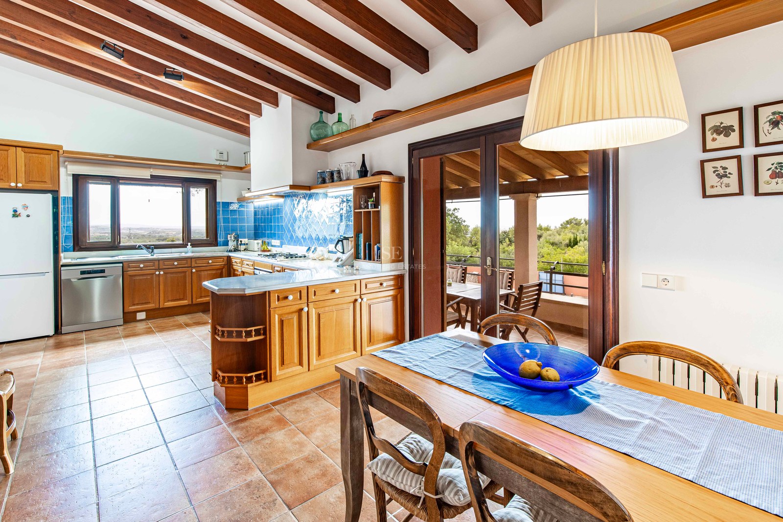 Cuisine avec meubles en bois, carrelage bleu, sol en terre cuite et poutres apparentes avec table à manger.