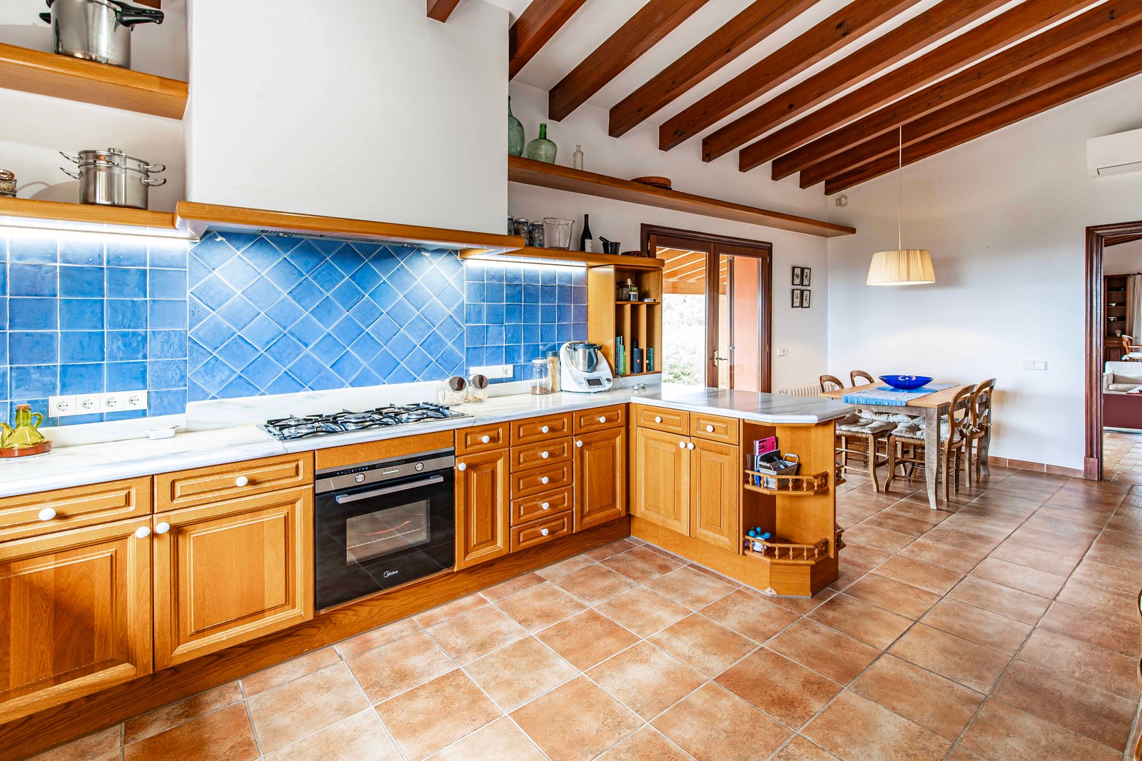 Cuisine rustique avec meubles en bois, crédence bleue, sol en terre cuite et plafond à poutres apparentes.