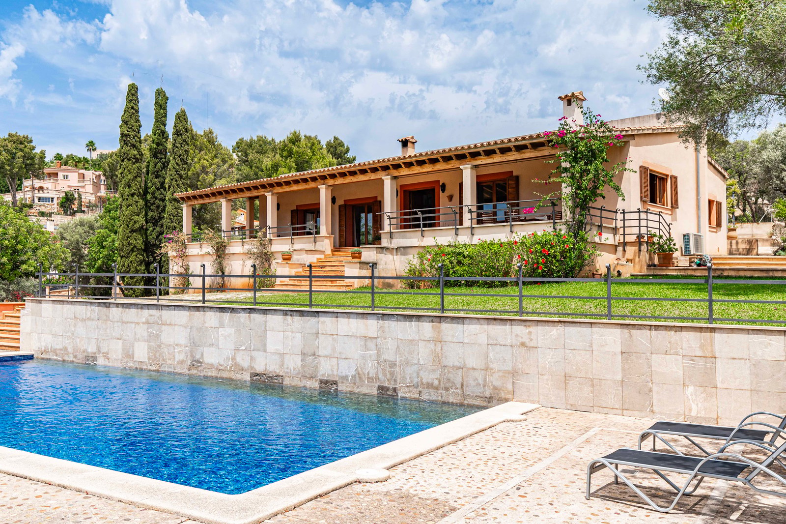 Grande piscine bleue et transats devant une villa méditerranéenne avec mur en pierre, pelouse et terrasse couverte.