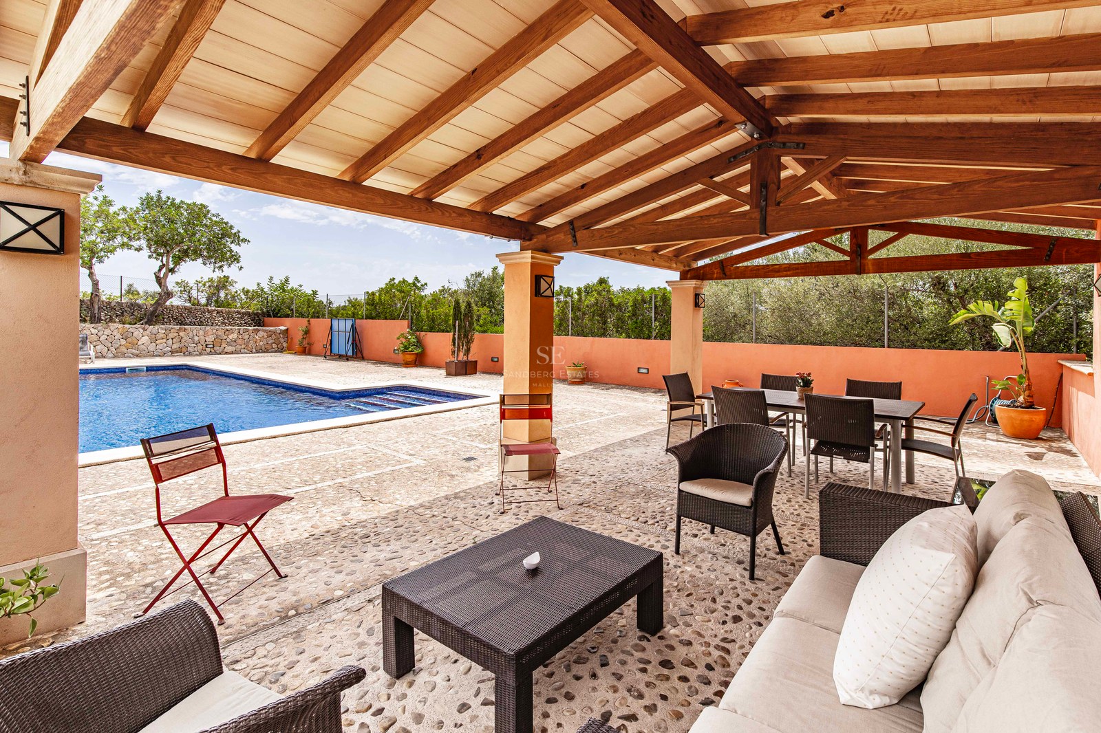 Terrasse couverte avec pergola en bois, sol en pierre naturelle, coin repas et salon face à une piscine bleue.