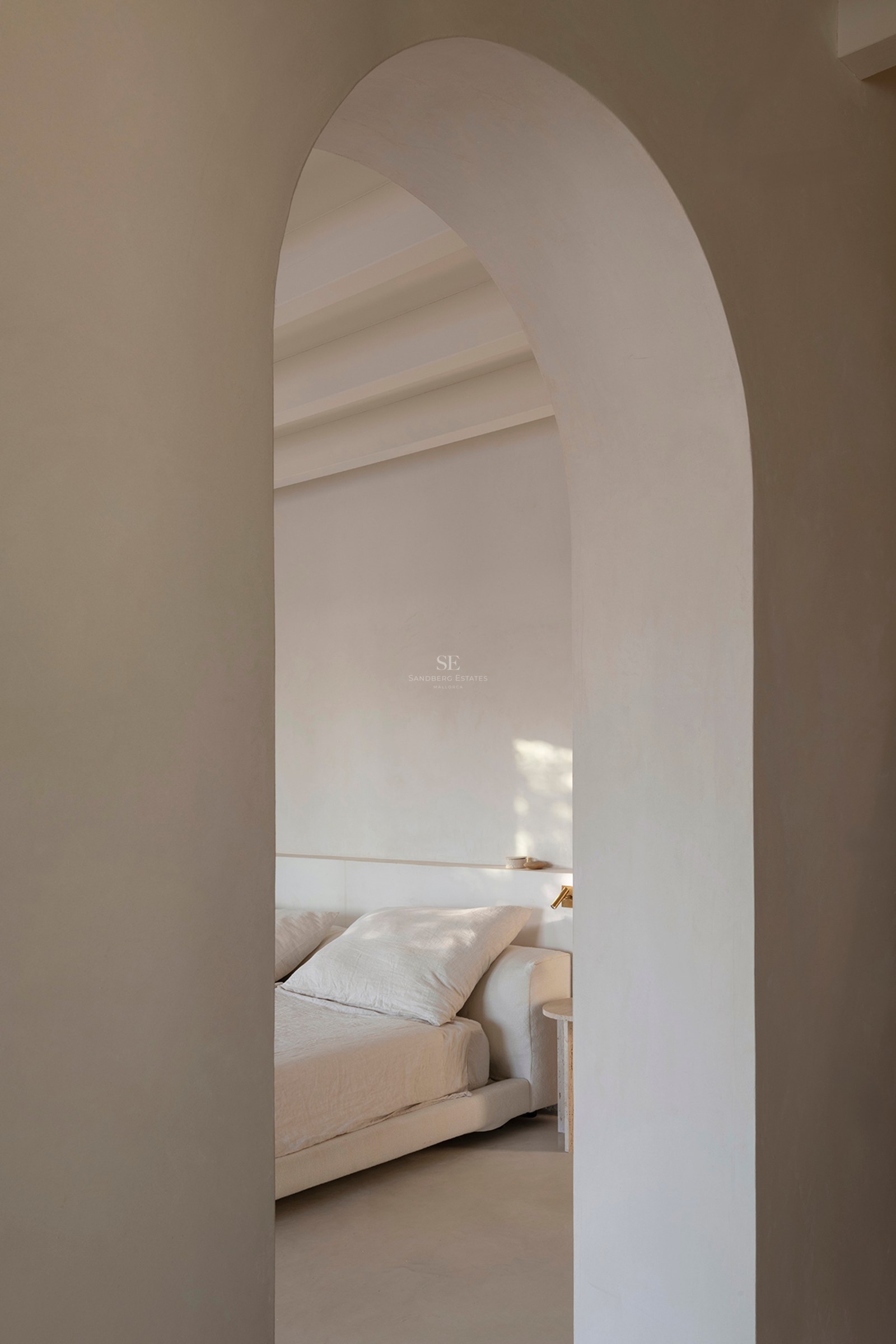 Minimalistisches Schlafzimmer mit Leinenbettwäsche, gesehen durch einen glatten Rundbogen in warmen Neutraltönen.