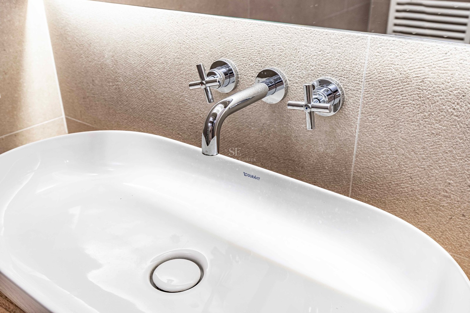 Närbild på ett vitt Duravit-tvättställ med väggmonterad kromad blandare mot en texturerad stenvägg.