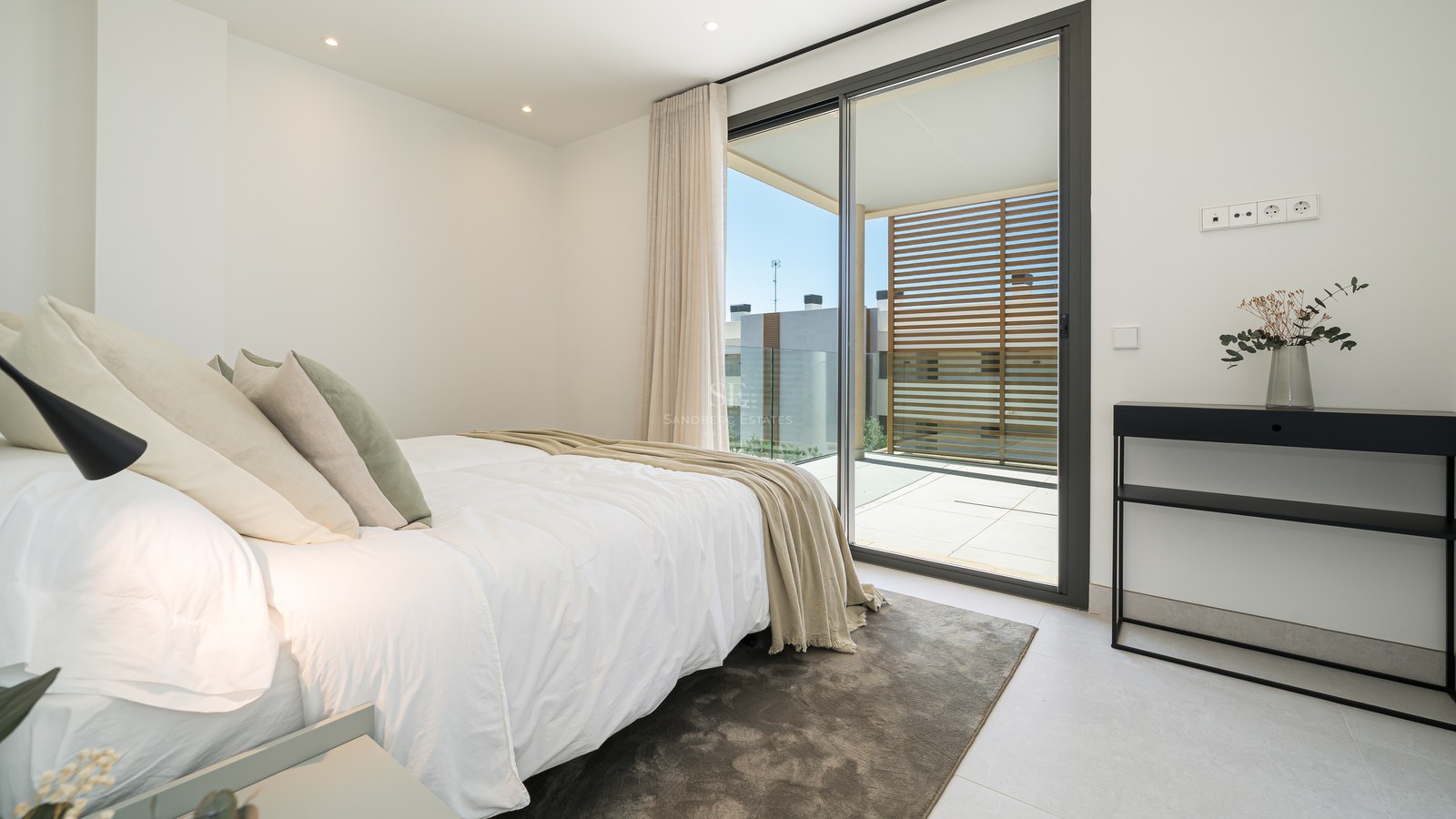 Contemporary bedroom featuring a white bed, black console table, and sliding glass doors leading to a sunny terrace.