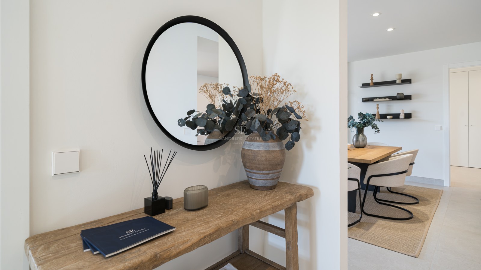 Minimalist entryway featuring a rustic wooden console table, round black mirror, and view into a modern dining room.