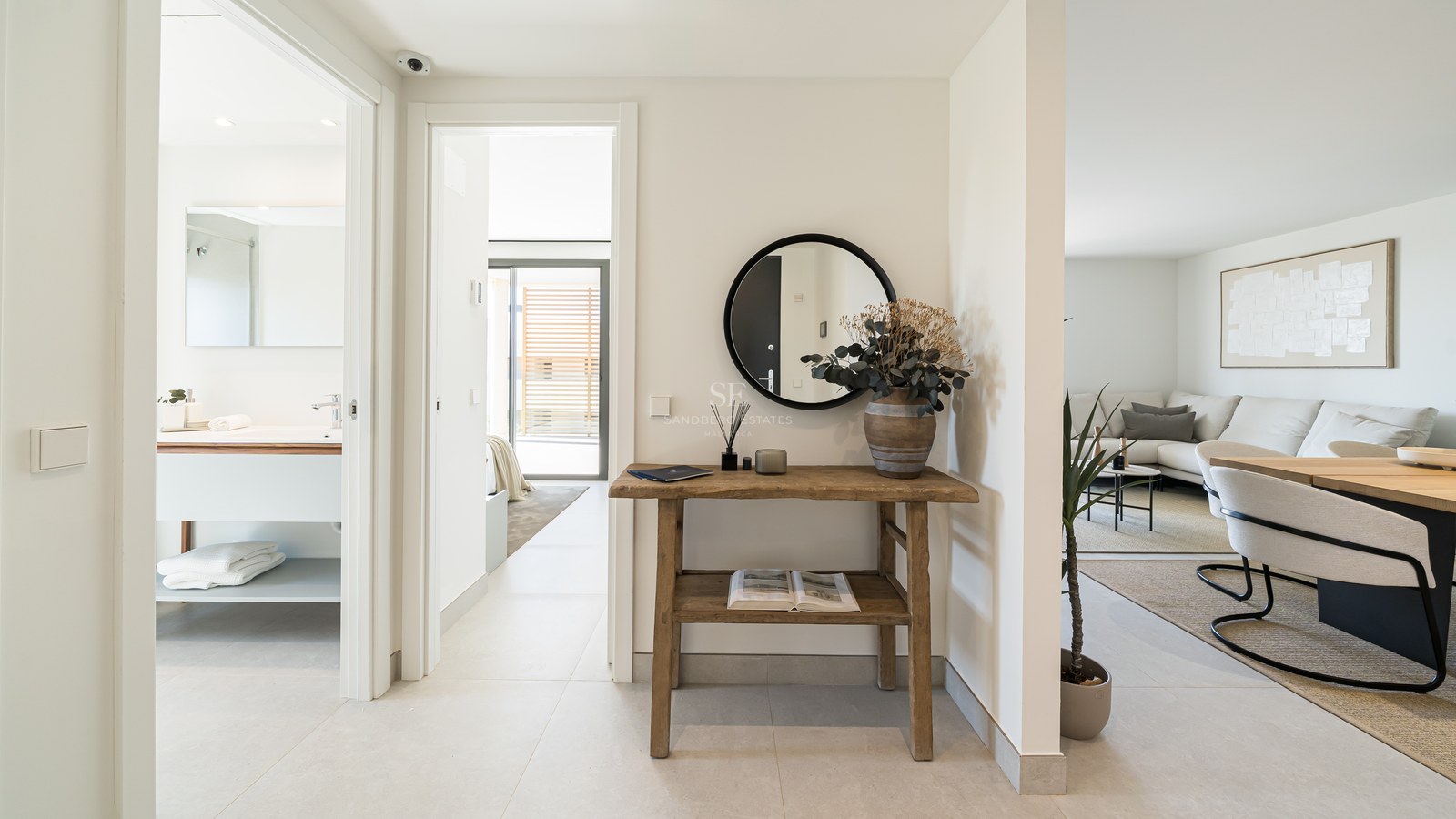 Bright hallway featuring a rustic wood console, round mirror, and views into a modern bathroom and living area.