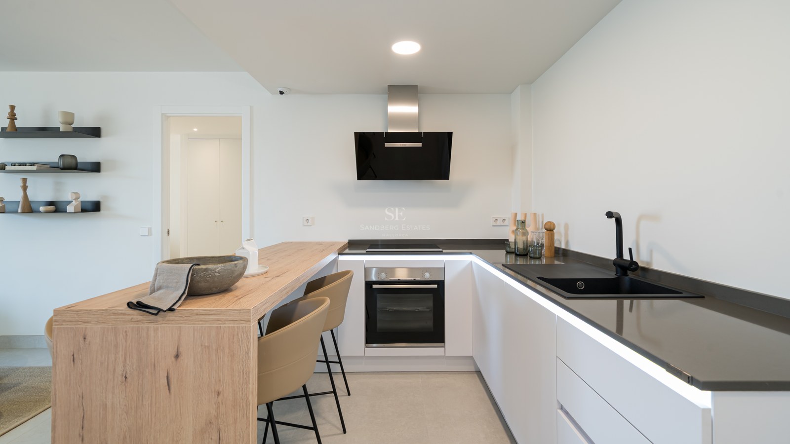 Minimalist white kitchen featuring a light oak breakfast bar, black sink, and sleek integrated appliances.