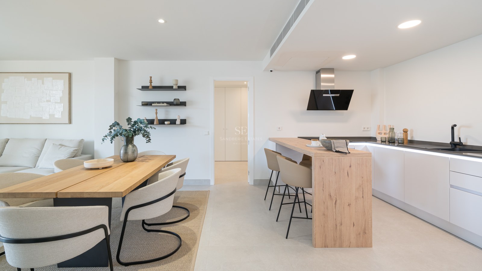 Minimalist white kitchen with wood island and adjacent dining area featuring designer chairs and light stone flooring.
