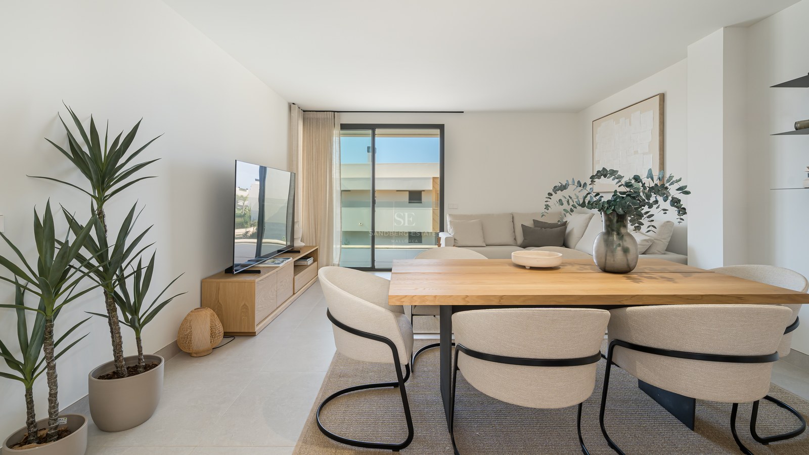 Bright open-plan room featuring a light wood dining table, cream chairs, a sectional sofa, and glass terrace doors.