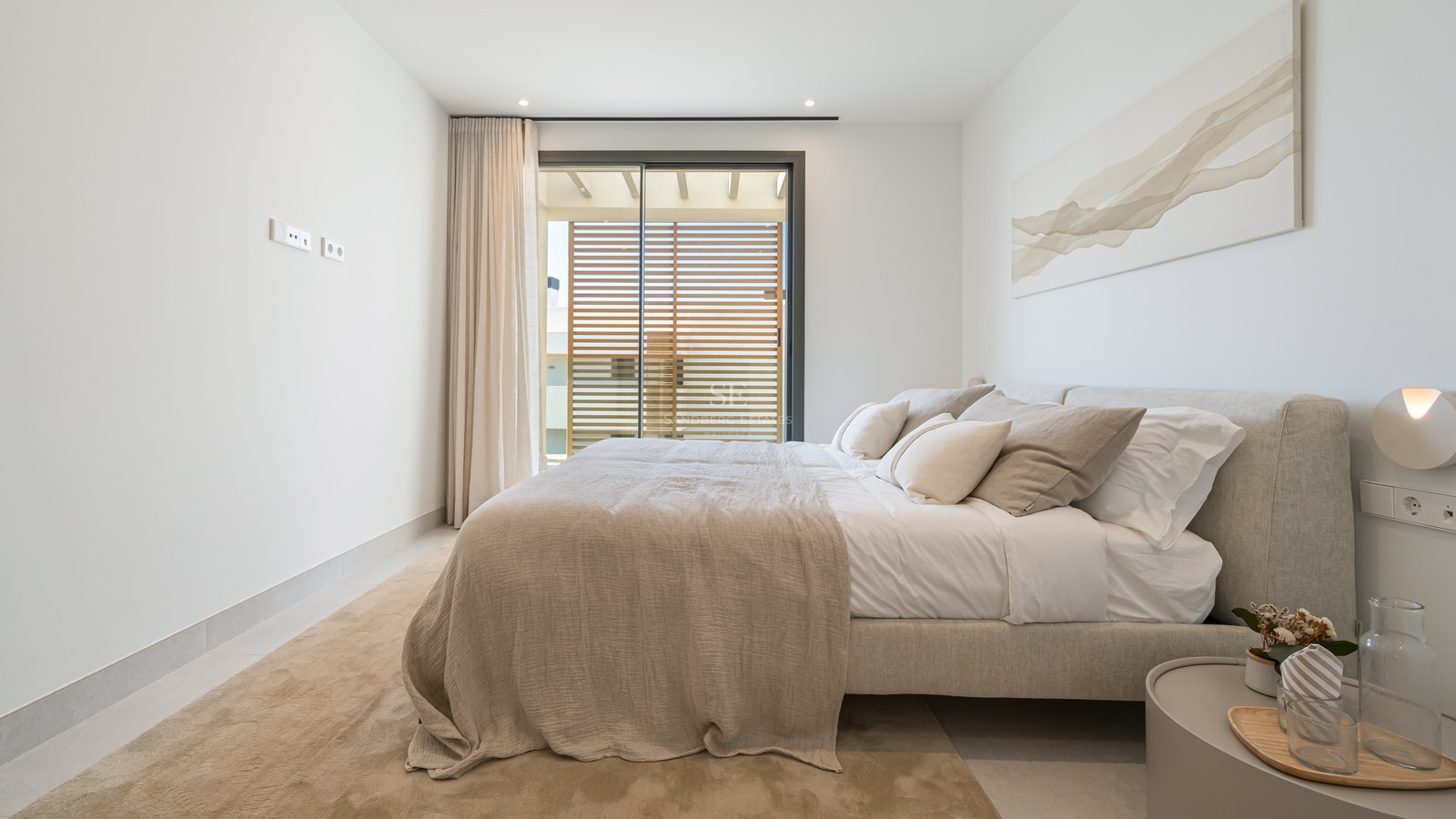 Bright modern bedroom with a beige upholstered bed, abstract wall art, and glass doors opening to a sunny terrace.