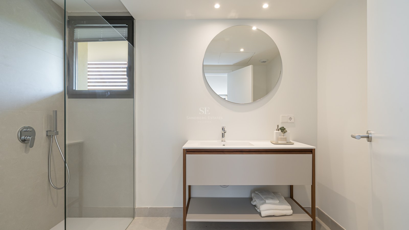 A modern bathroom featuring a walk-in glass shower, a circular mirror over a wooden vanity, and grey tiled walls.