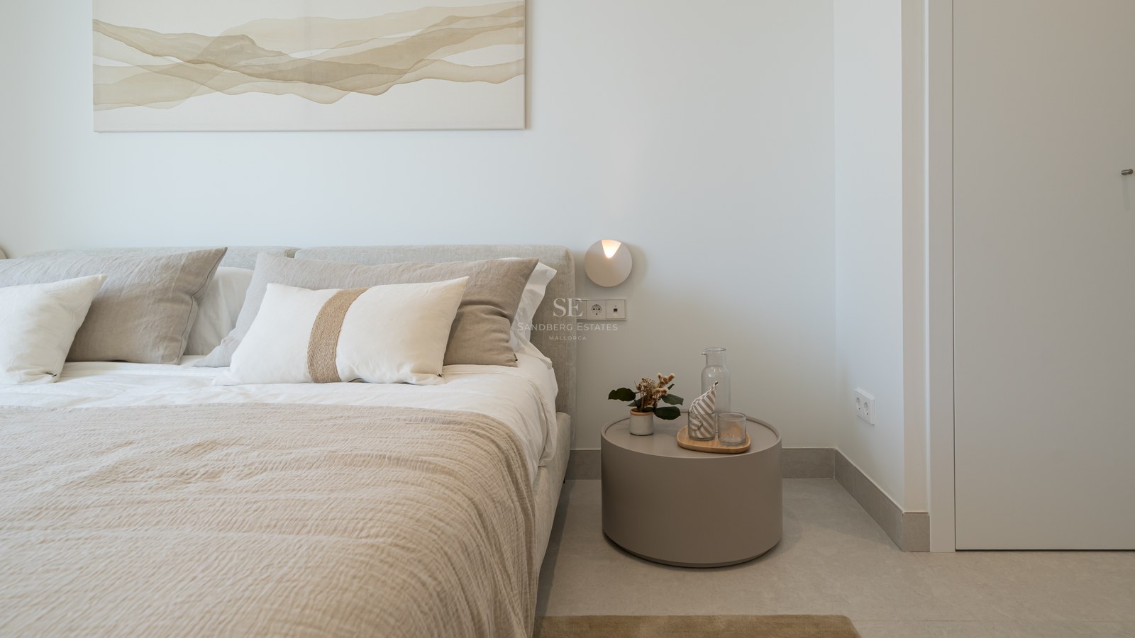 Close-up of a bed with beige linen bedding next to a round minimalist nightstand and abstract wall art.