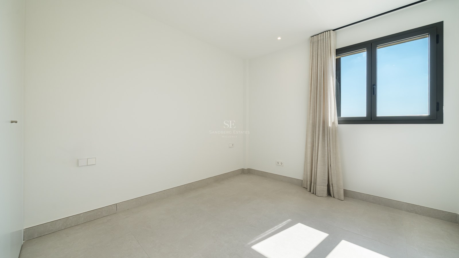 An empty minimalist room with white walls, large gray floor tiles, and a window with neutral curtains.