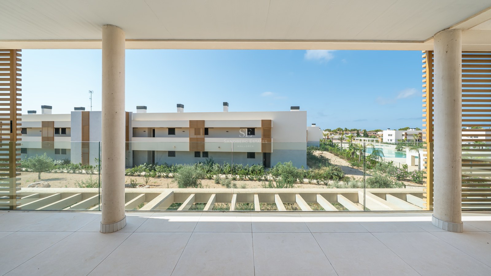 A spacious covered terrace with glass balustrades and concrete pillars overlooking modern apartments and a pool.