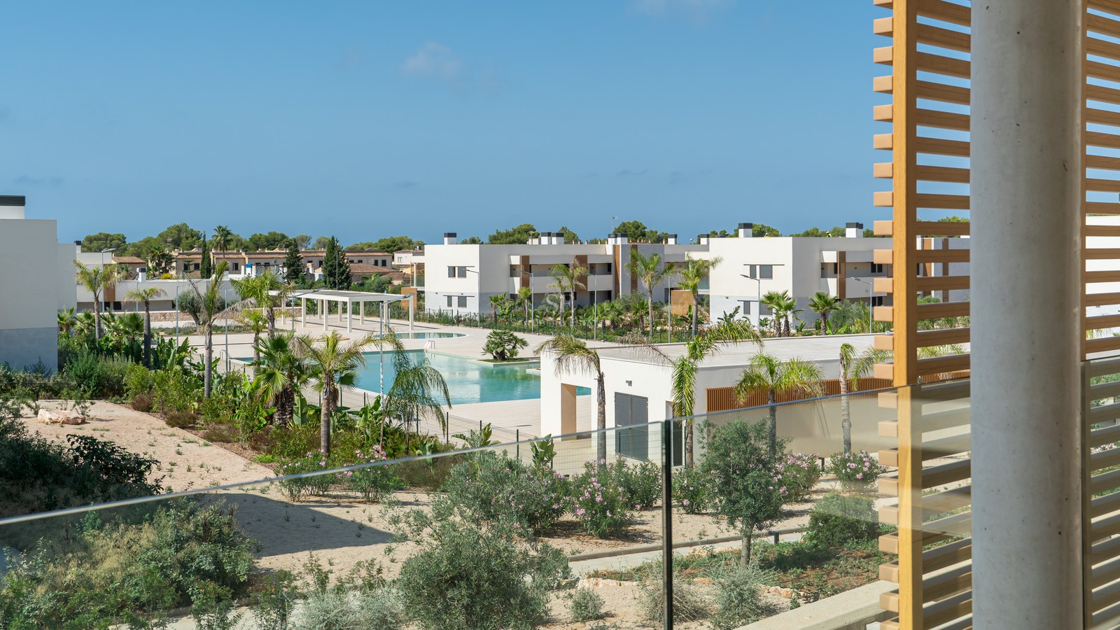 View from a modern terrace with glass railing overlooking a large turquoise swimming pool, palm trees, and white buildings.