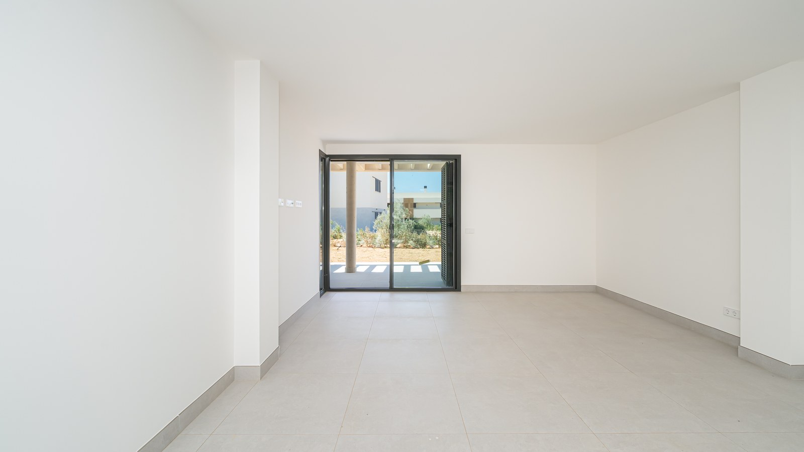 Empty modern room with white walls, large grey floor tiles, and a sliding glass door opening to a garden.