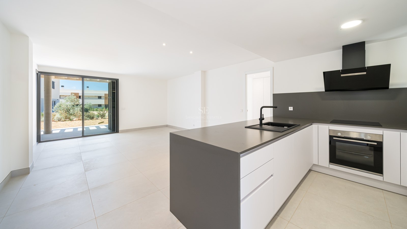 Minimalist white kitchen featuring a grey island, black appliances, and large sliding glass doors leading outside.