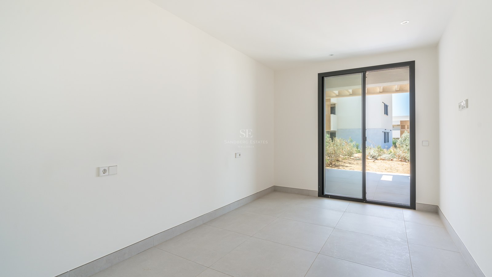 Empty modern bedroom with white walls, grey tile flooring, and sliding glass door opening to a private garden patio.