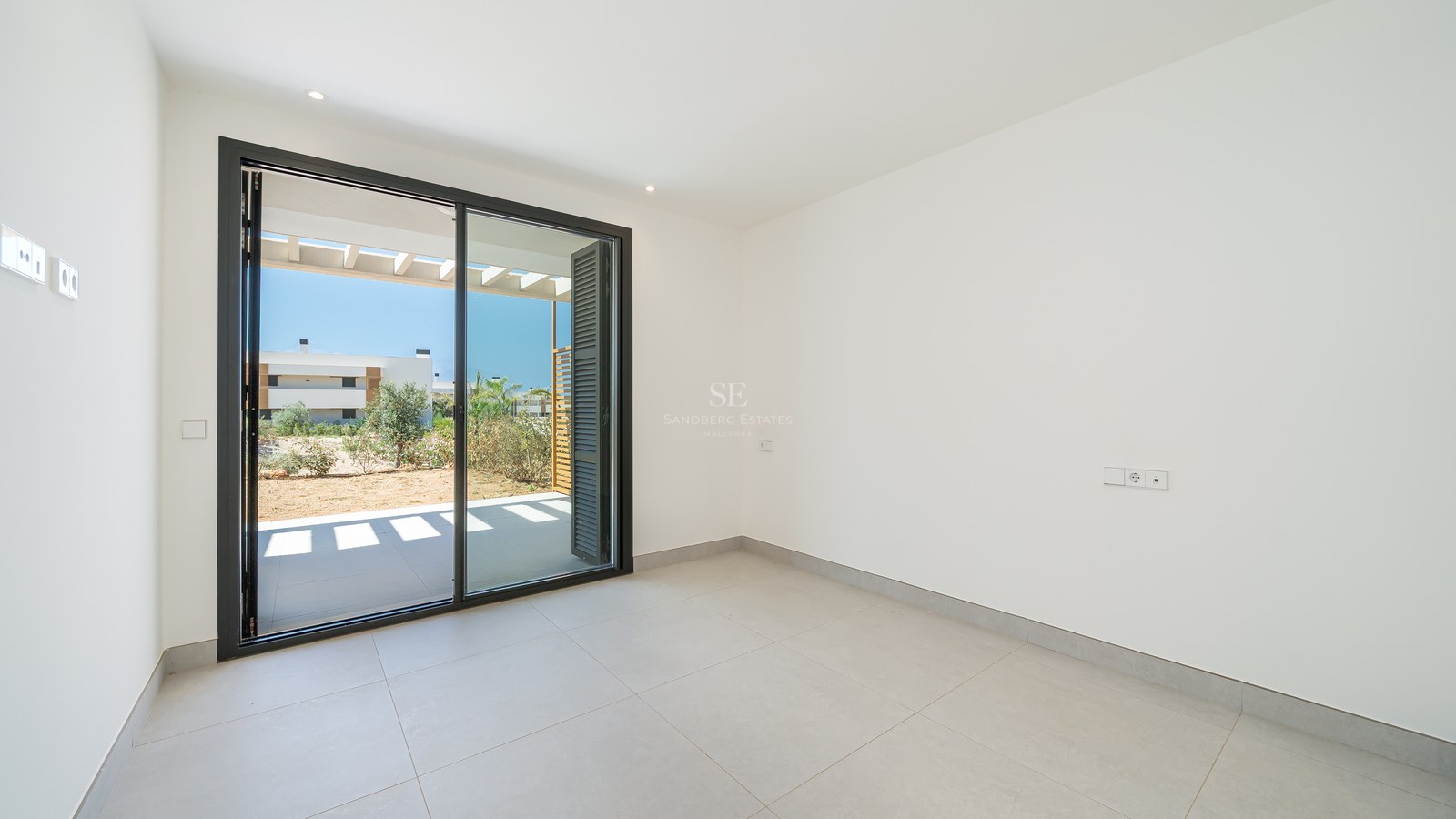 Empty modern room with white walls, grey tiled flooring, and a large sliding glass door opening onto a sunlit terrace.