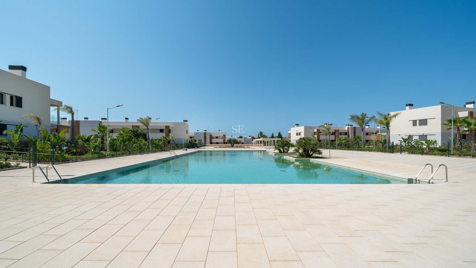 Large turquoise swimming pool surrounded by light stone paving and modern white villas under a clear blue sky.