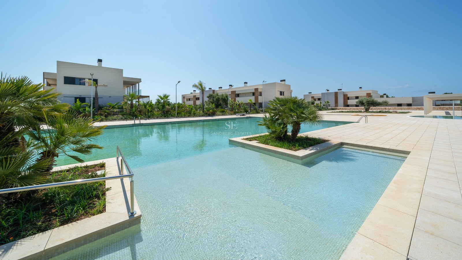 Spacious turquoise swimming pool surrounded by stone tiling and contemporary white buildings under a clear blue sky.