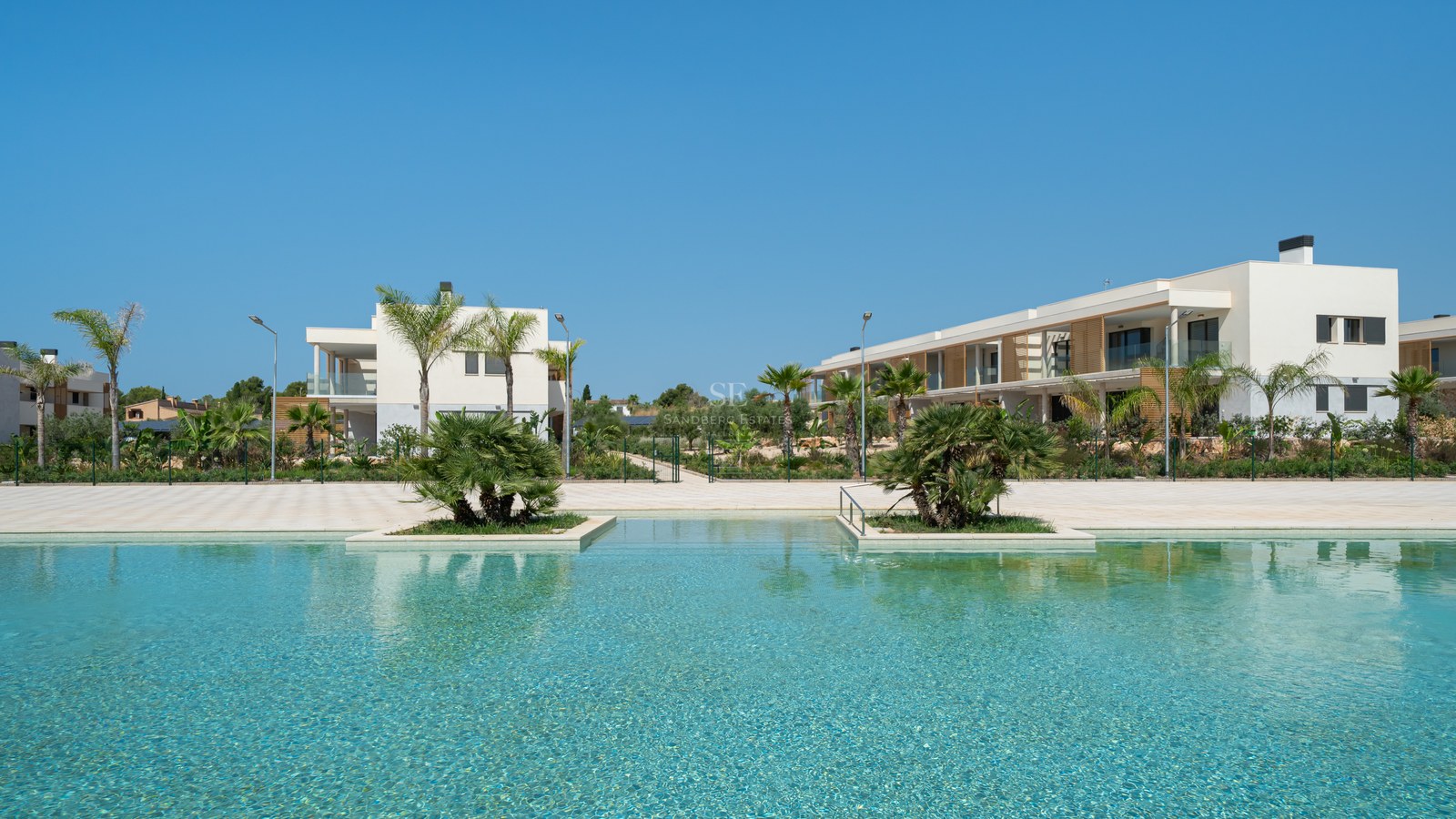 Large turquoise swimming pool with palm tree islands in front of white modern Mediterranean buildings under a blue sky.