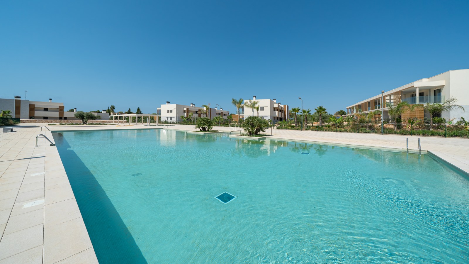 Large turquoise outdoor pool surrounded by cream stone tiles and modern white apartment buildings under a clear blue sky.