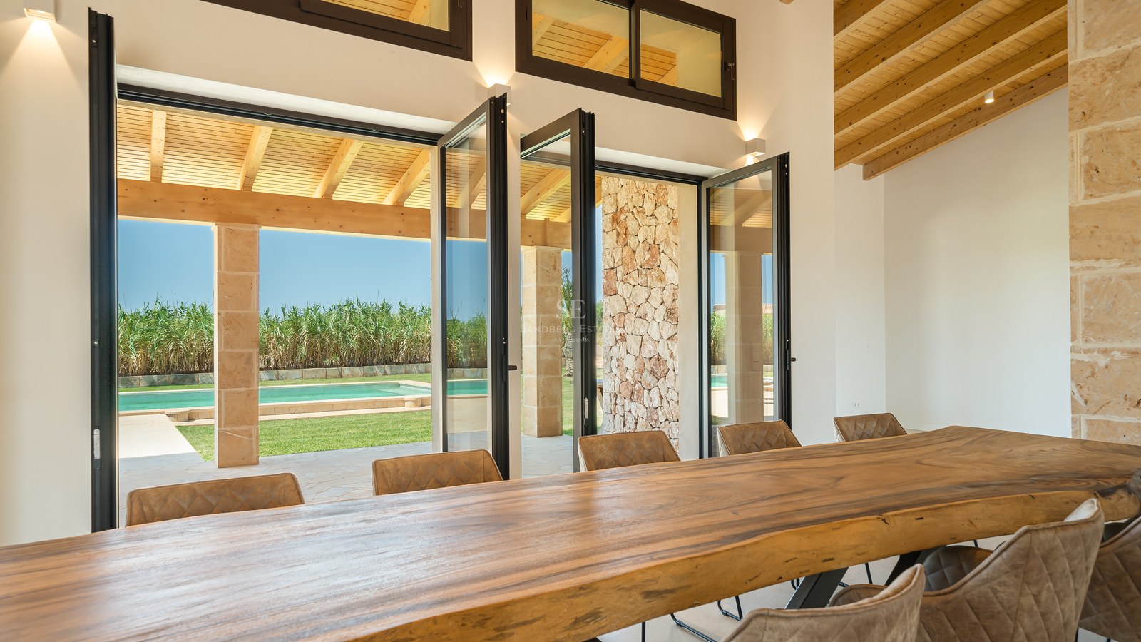 Grande table de salle à manger en bois naturel face à des baies vitrées s'ouvrant sur le jardin et la piscine.