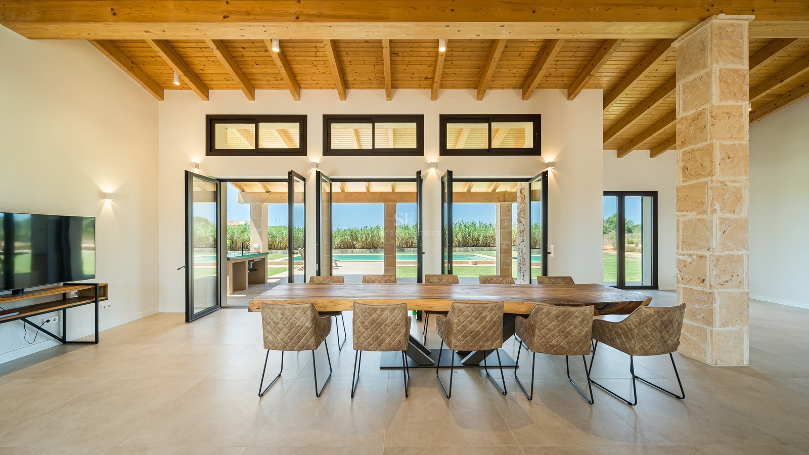 Grande salle à manger avec table en bois massif, plafond à poutres et pilier en pierre naturelle face à la piscine.