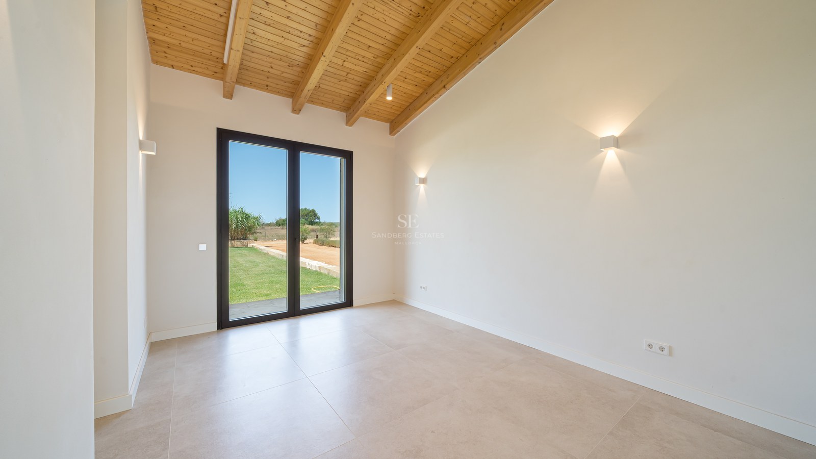 Pièce moderne vide avec plafond voûté en poutres de bois, sol en carrelage beige et porte vitrée sur le jardin.