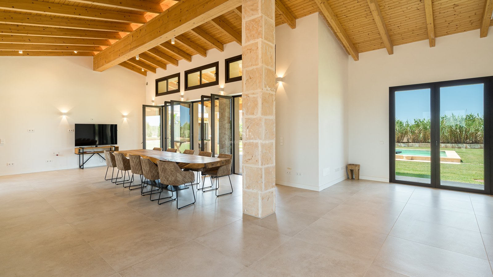 Grande salle à manger avec table en bois massif, colonne en pierre, plafond à poutres et vue sur le jardin.