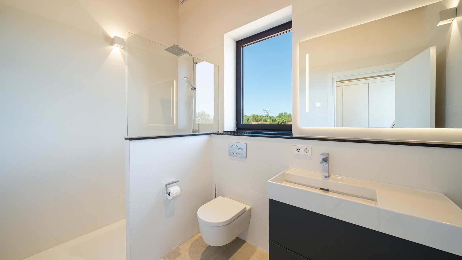Salle de bains blanche minimaliste avec douche en verre, WC suspendus et meuble vasque sombre sous une fenêtre.