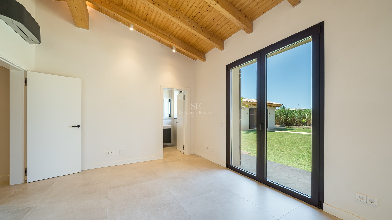 Chambre vide avec de hauts plafonds en poutres de bois et des baies vitrées donnant sur un jardin ensoleillé.