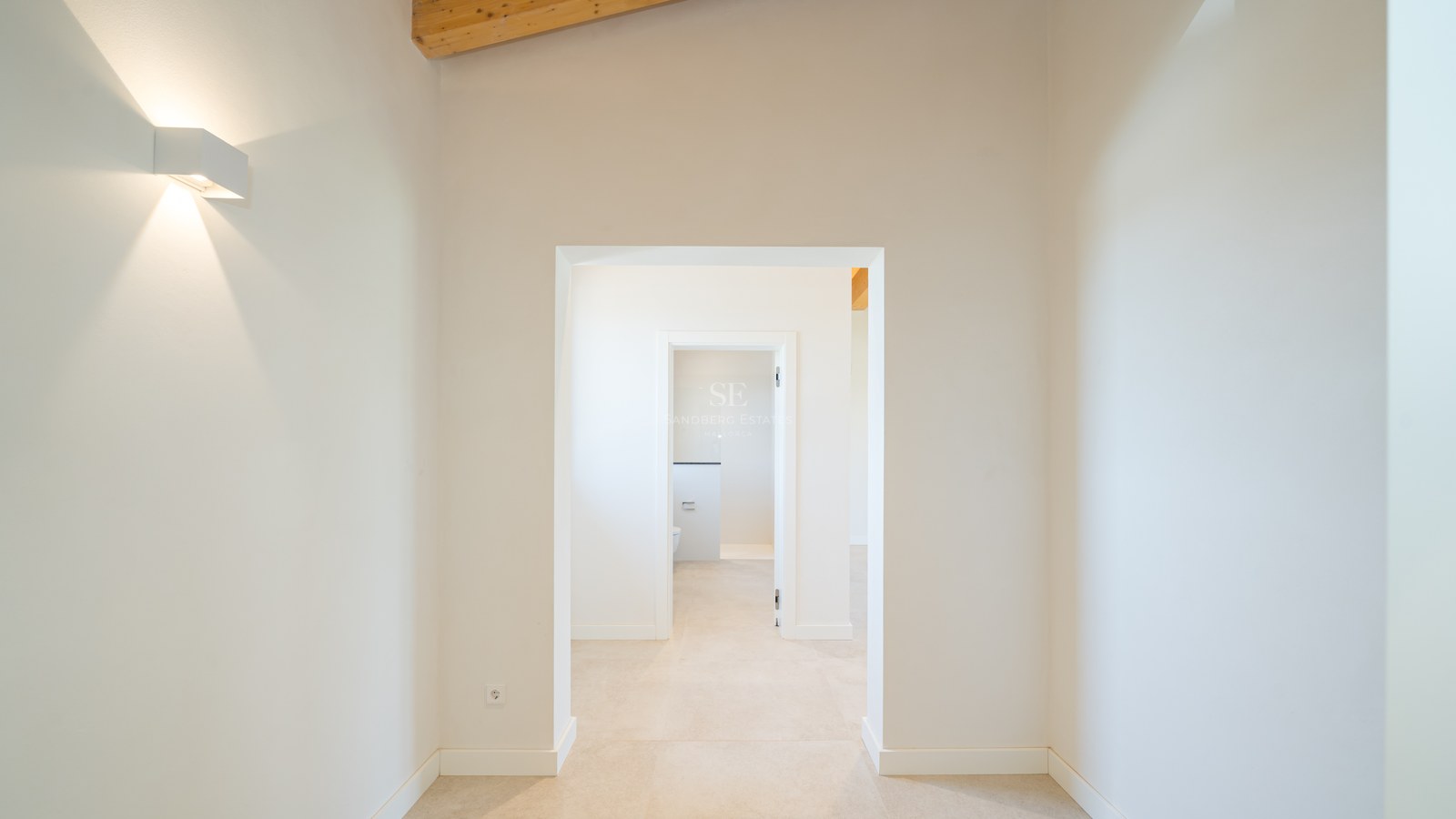 Couloir intérieur lumineux avec murs blancs, sol en pierre et hauts plafonds avec poutres en bois.