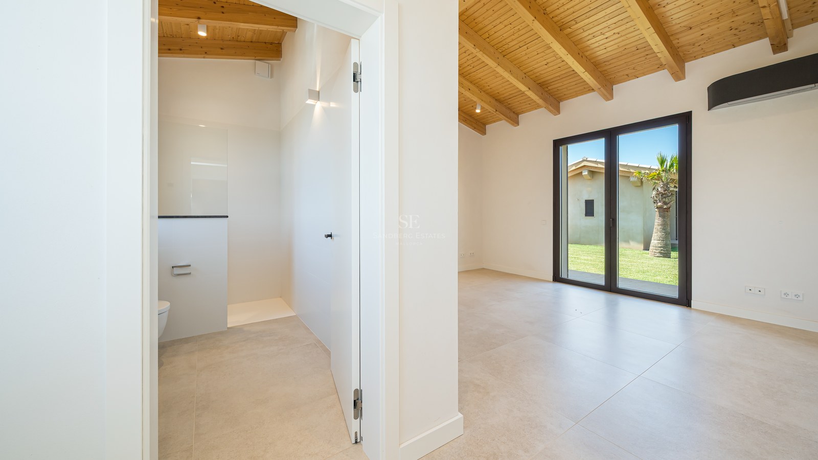 Chambre lumineuse avec poutres en bois clair, carrelage grand format et aperçu de la salle de bain attenante.