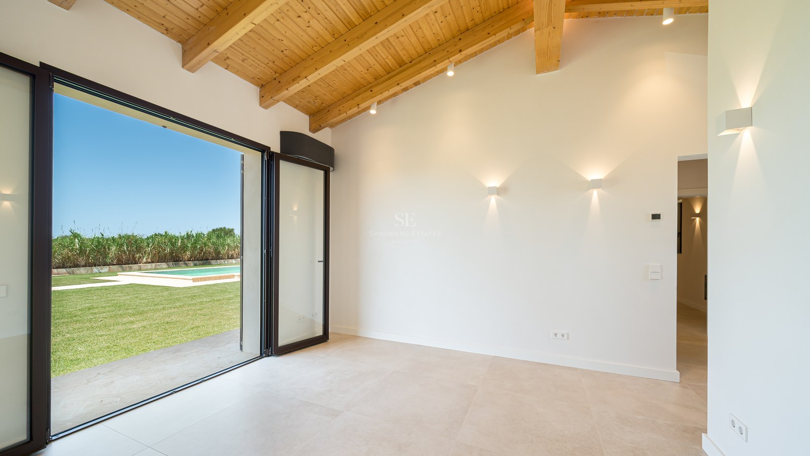 Pièce vide avec de hauts plafonds voûtés en bois et de grandes baies vitrées ouvrant sur une pelouse et une piscine.