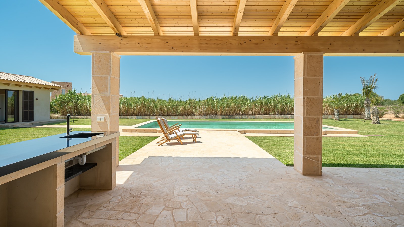 Terrasse en pierre avec cuisine d'été donnant sur une piscine et un jardin sous un ciel bleu.