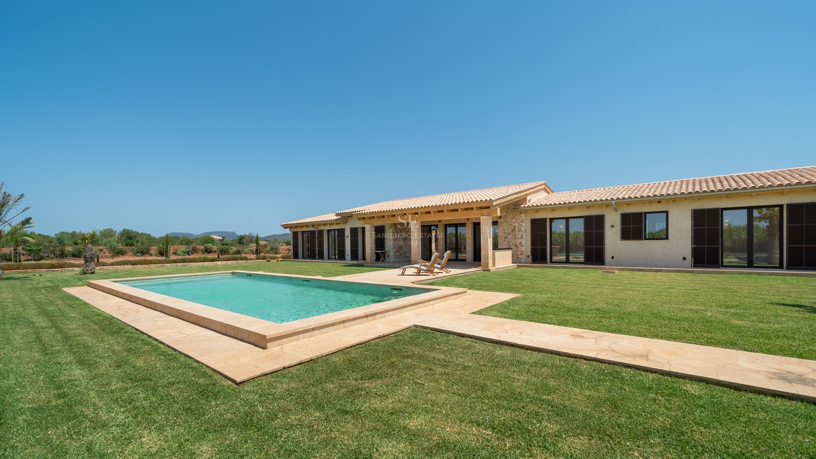 Piscine rectangulaire sur une grande pelouse devant une villa moderne en pierre sous un ciel bleu.