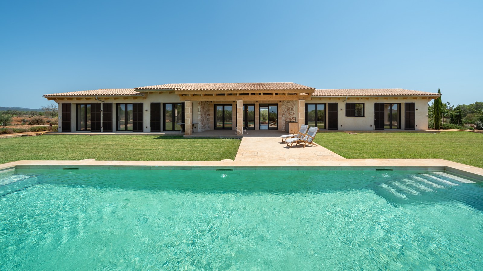 Piscine rectangulaire turquoise devant une villa moderne en pierre de plain-pied avec volets sombres et pelouse.