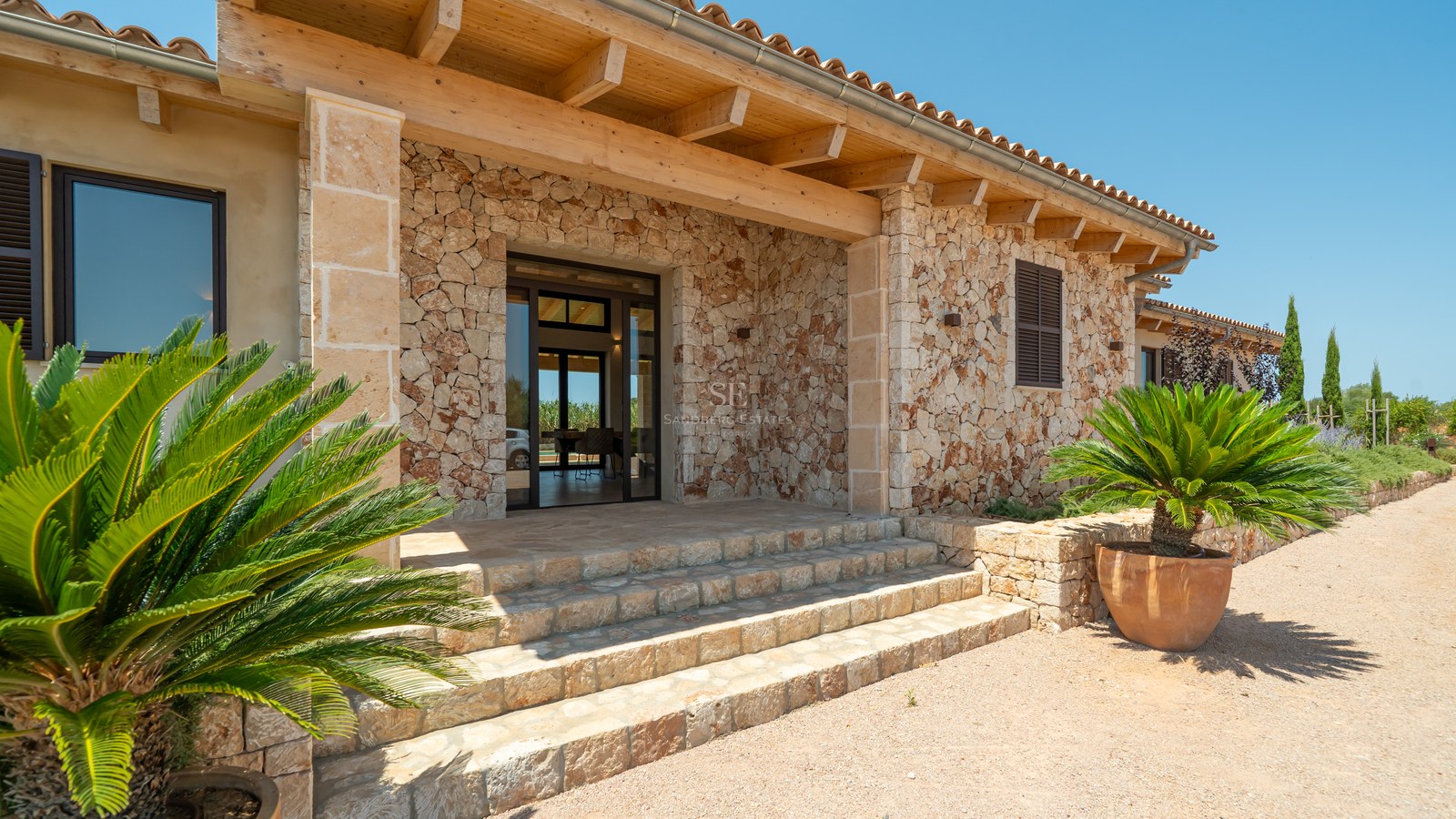 Façade en pierre naturelle avec poutres en bois, escaliers en pierre et plantes exotiques sous un soleil radieux.