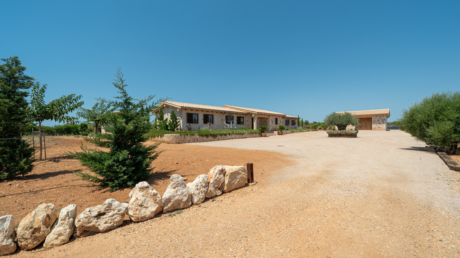 Façade d'une villa méditerranéenne avec murs en pierre, toit en terre cuite et allée de gravier sous un ciel bleu.