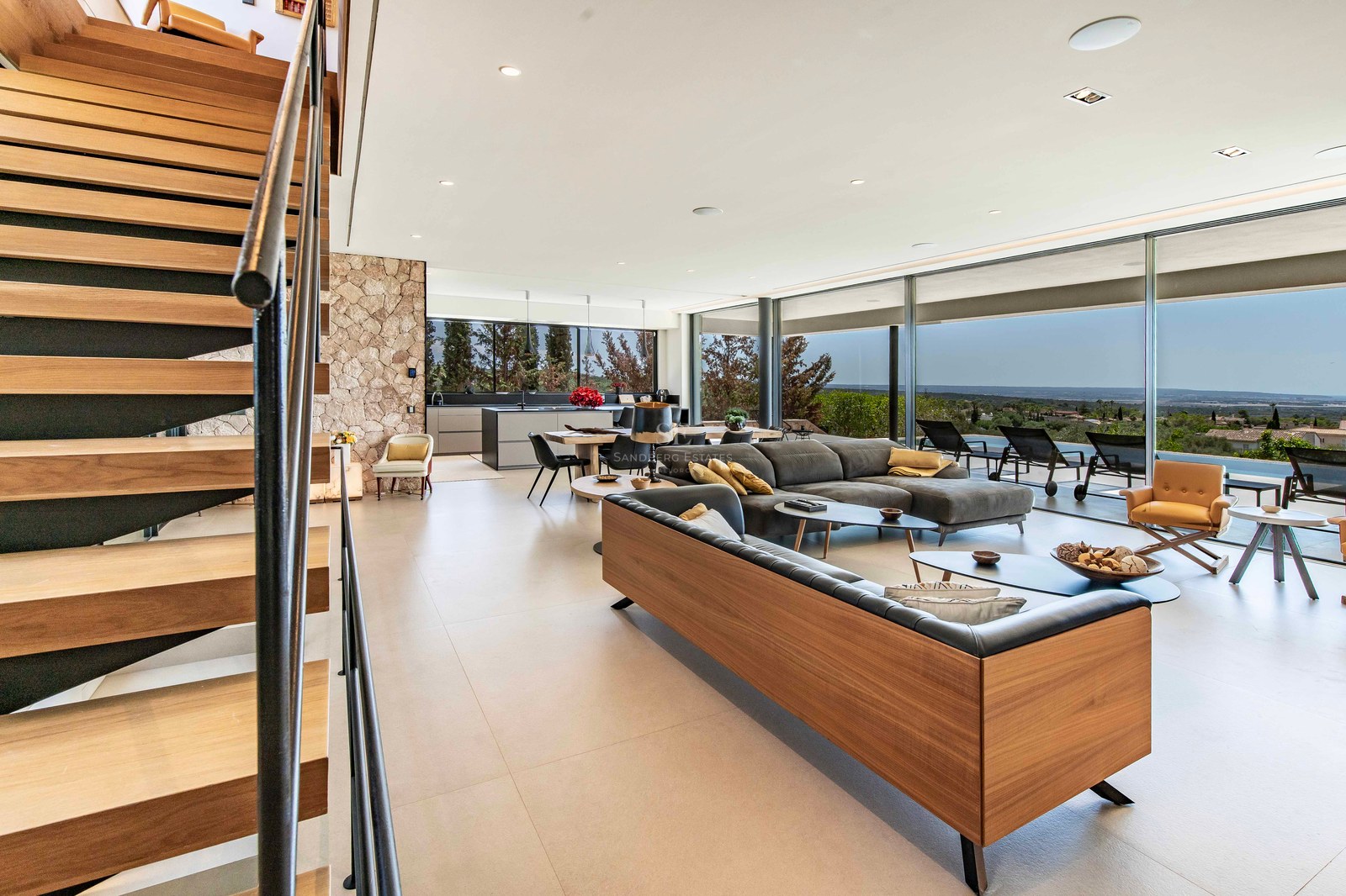 Spacious modern living room with wooden stairs, stone wall, and floor-to-ceiling windows overlooking a terrace and landscape.