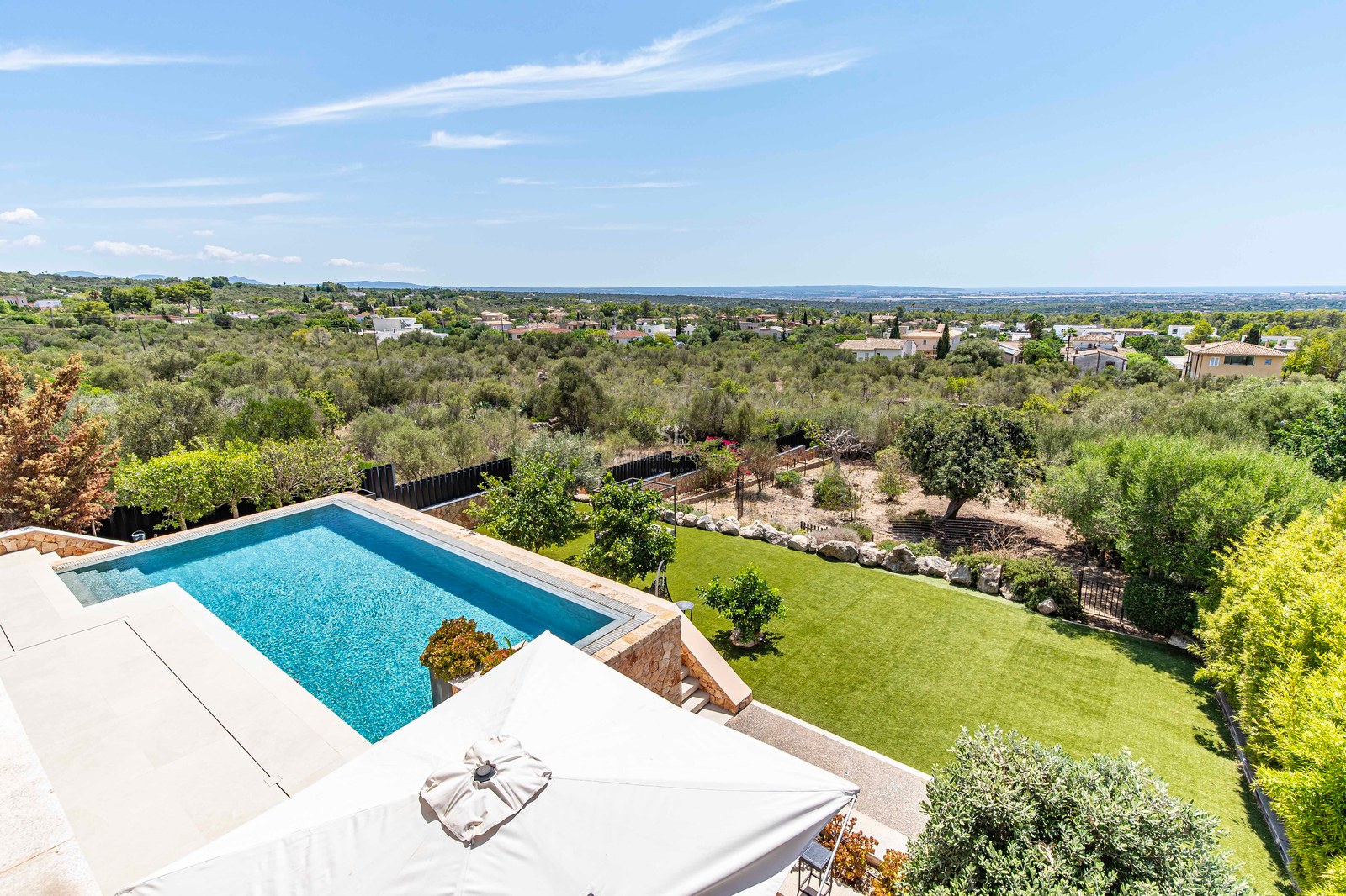 Elevated view of a rectangular blue swimming pool surrounded by a natural stone terrace and lawn overlooking the sea.