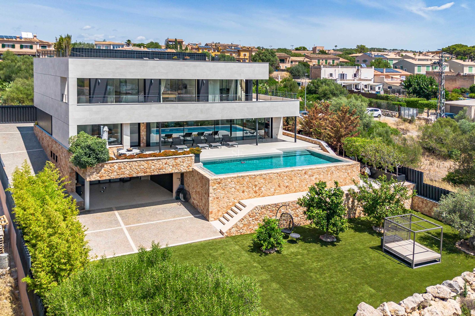 Elevated view of a three-story contemporary villa with stone walls, glass balconies, swimming pool, and green lawn.